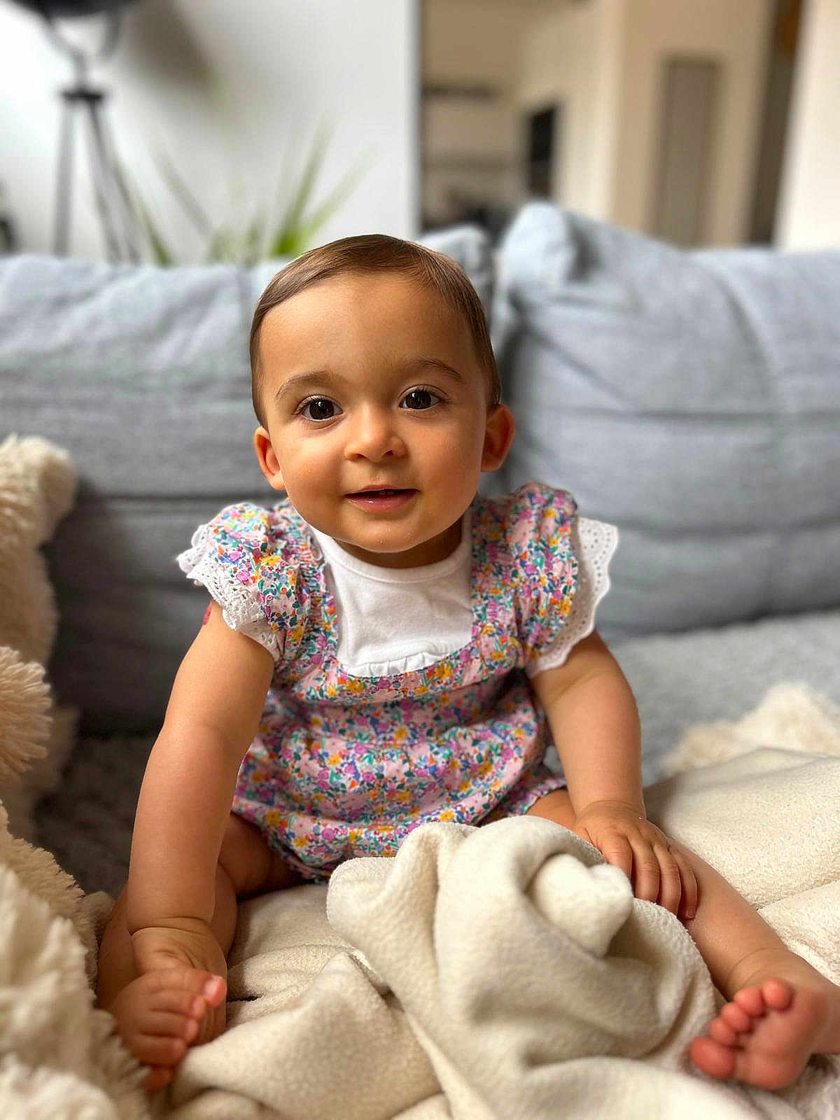 Ava a rejoint le concours — aidez-le/la à gagner de superbes lots ! baby, child, infant, sitting, couch, blanket, smiling, face, cute, indoors, soft, floral_dress, cozy, happy, person, young, skin, foot, hand, portrait