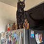 cat, black_cat, yellow_eyes, refrigerator, magnets, kitchen, indoor, appliance, cabinet, skeleton_decor, golden_statue, pet, curious, standing, fridge_top, ceiling, photograph, decor, metal_surface, portrait_photos
