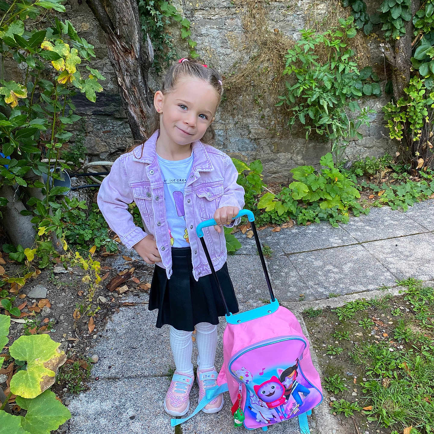 Emili a rejoint le concours — aidez-le/la à gagner de superbes lots ! backpack, black_skirt, child, cute, daylight, garden, girl, greenery, nature, outdoor, pathway, playful, portrait, posing, purple_jacket, smiling, sneakers, stone_wall, white_tights, young