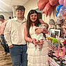 baby, balloons, celebration, couple, decorations, family, female, gift_bags, indoor, male, newborn, party, people, photo_frames, smile, standing, wooden_floor, white_dress, jeans, pacifier