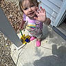 child, toddler, smiling, waving, outdoor, porch, driveway, house, concrete, gravel, toy, toy_vehicle, pink_clogs, overalls, purple_shirt, blonde_hair, blue_eyes, portrait, daylight, happy