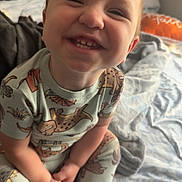 Kal-El joined the competition — help win amazing prizes! bed, bedroom, blanket, casual_clothing, child, cozy, cute, dinosaur_print, face, happy, indoors, pajamas, person, sitting, small_object, smiling, soft_lighting, toddler, toy, young_child