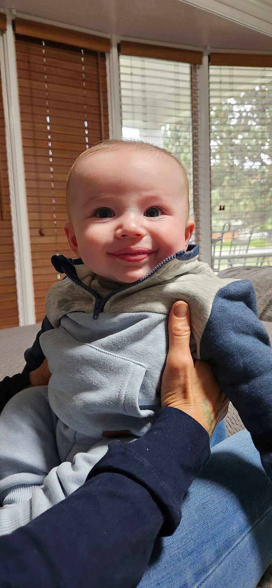 Declan is registered to the contest to win money with this photo: baby, child, smile, face, person, indoor, window, blinds, hand, clothing, hoodie, jeans, sitting, happy, infant, portrait, light, adult_hand, cozy, casual
