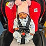 baby, sleeping, car_seat, pink_hat, pom_poms, blue_onesie, star_pattern, infant, child, safety_harness, red_car_seat, black_straps, cozy_clothing, indoors, peaceful, cute, newborn, headwear, toddler, seatbelt