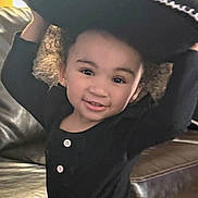 Caliyah is registered to the contest to win money with this photo: toddler, child, smiling, curly_hair, black_clothing, hat, indoor, couch, leather, happy, portrait, cute, playful, face, person, young_child, buttoned_shirt, home, childhood, joy