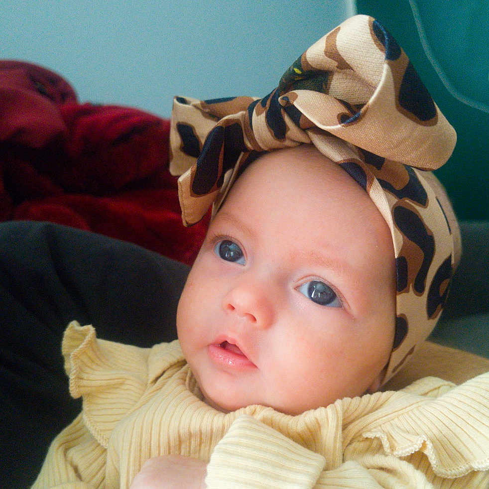 Anaya is registered to the contest to win money with this photo: baby, child, infant, portrait, face, eyes, wide_eyed, looking_at_camera, bonnet, camouflage, hat, yellow_clothing, blanket, indoor, close_up, headshot, couch, red_fabric, soft_lighting, adorable