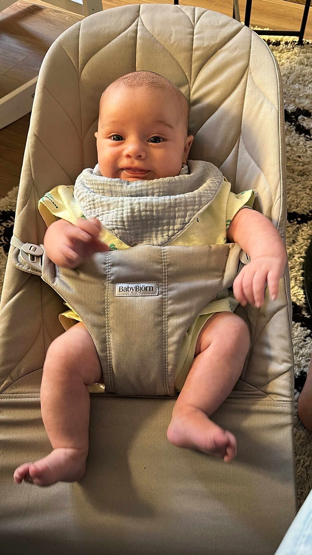 Alessio a rejoint le concours — aidez-le/la à gagner de superbes lots ! baby, infant, bouncer, babybjorn, bib, yellow_clothing, smile, happy, cute, indoors, carpet, wooden_floor, baby_seat, child, person, legs, arms, face, skin, comfort