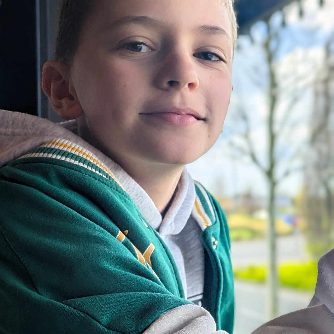 Antoine participe au concours pour gagner de l'argent avec cette photo : child, boy, portrait, smile, casual, window, indoor, clothing, hoodie, green_jacket, face, young, person, daylight, background, trees, outdoor_blur, expression, head, leisure