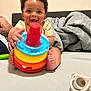 baby, child, smiling, toy, colorful, stackable_toy, pacifier, bed, blanket, curly_hair, happy, indoors, person, infant, playful, tattoo, hand, foot, face, expression