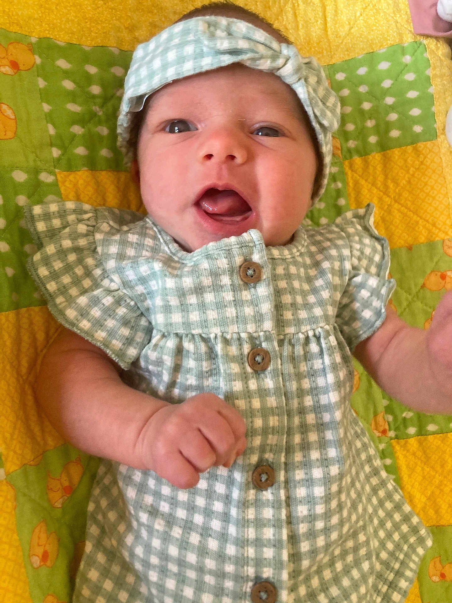 Raegan is registered to the contest to win money with this photo: baby, newborn, infant, headband, dress, gingham, buttons, blanket, quilt, open_mouth, smiling, face, eyes, hand, arm, lying_down, portrait, green, yellow, cute