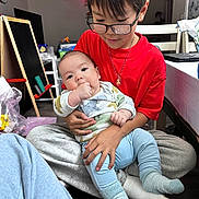 Liam joined the competition — help win amazing prizes! baby, child, glasses, red_shirt, blue_pants, socks, indoor, home, holding, affection, seated, table, chair, floor, toy, blackboard, necklace, face, person, family