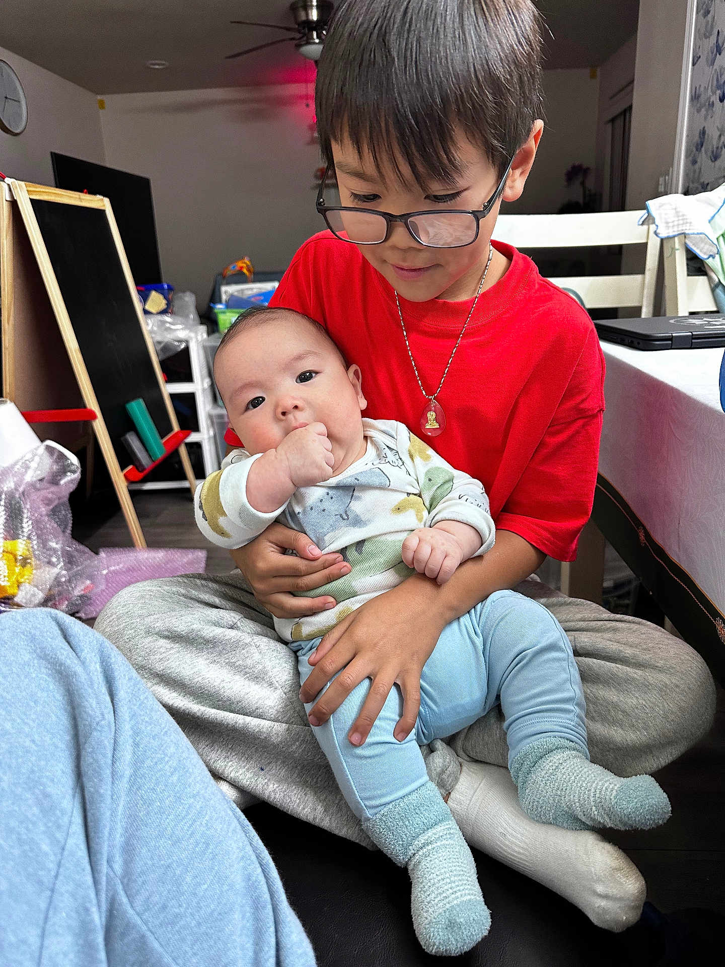 Liam joined the competition — help win amazing prizes! baby, child, glasses, red_shirt, blue_pants, socks, indoor, home, holding, affection, seated, table, chair, floor, toy, blackboard, necklace, face, person, family