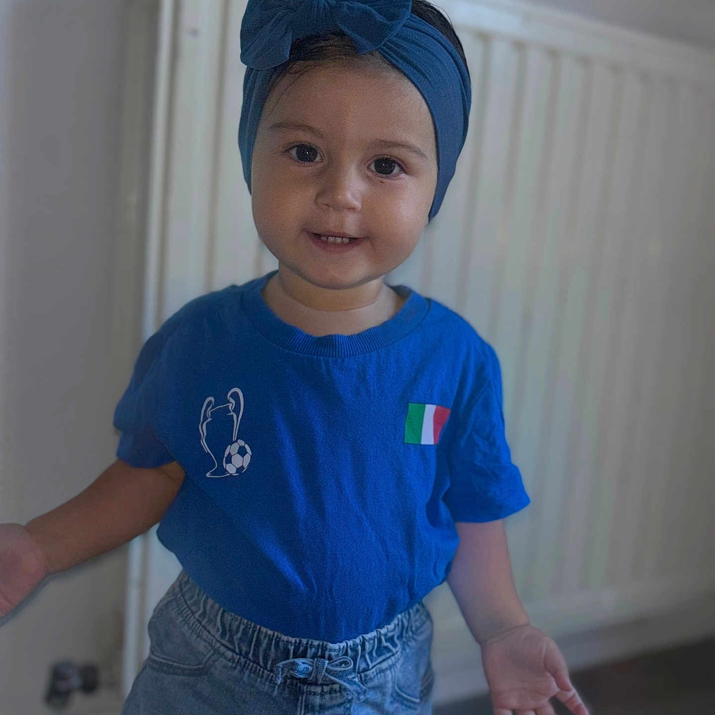Inaya a rejoint le concours — aidez-le/la à gagner de superbes lots ! blue_headband, blue_shirt, bow, casual_clothing, child, curious_expression, denim_shorts, floor, hands_open, indoor, italian_flag, portrait, radiator, short_hair, smiling, soccer_trophy, standing, toddler, wall, young_child