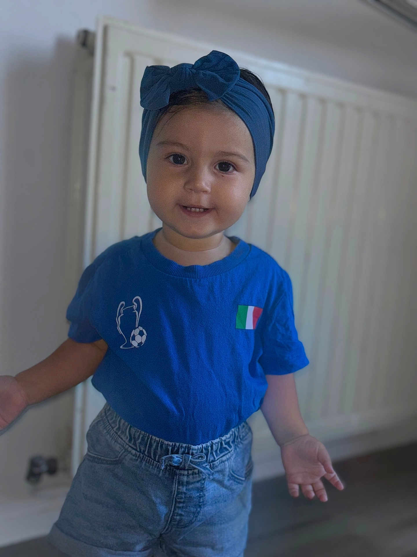 Inaya a rejoint le concours — aidez-le/la à gagner de superbes lots ! toddler, child, blue_headband, bow, blue_shirt, italian_flag, soccer_trophy, denim_shorts, indoor, radiator, curious_expression, standing, hands_open, short_hair, young_child, portrait, casual_clothing, floor, wall, smiling