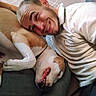 man, dog, pet, owner, cuddle, hugging, smiling, grinning, portrait, selfie, indoors, couch, sitting, white_shirt, collar, short_hair, beard, tile_floor, close_up, companionship