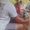 dog, boat, boating, captain, life_jacket, pink_clothing, human, person, steering_wheel, cockpit, watercraft, outdoor, nature, trees, window, travel, phone_screenshot, text_overlay, summer, safety