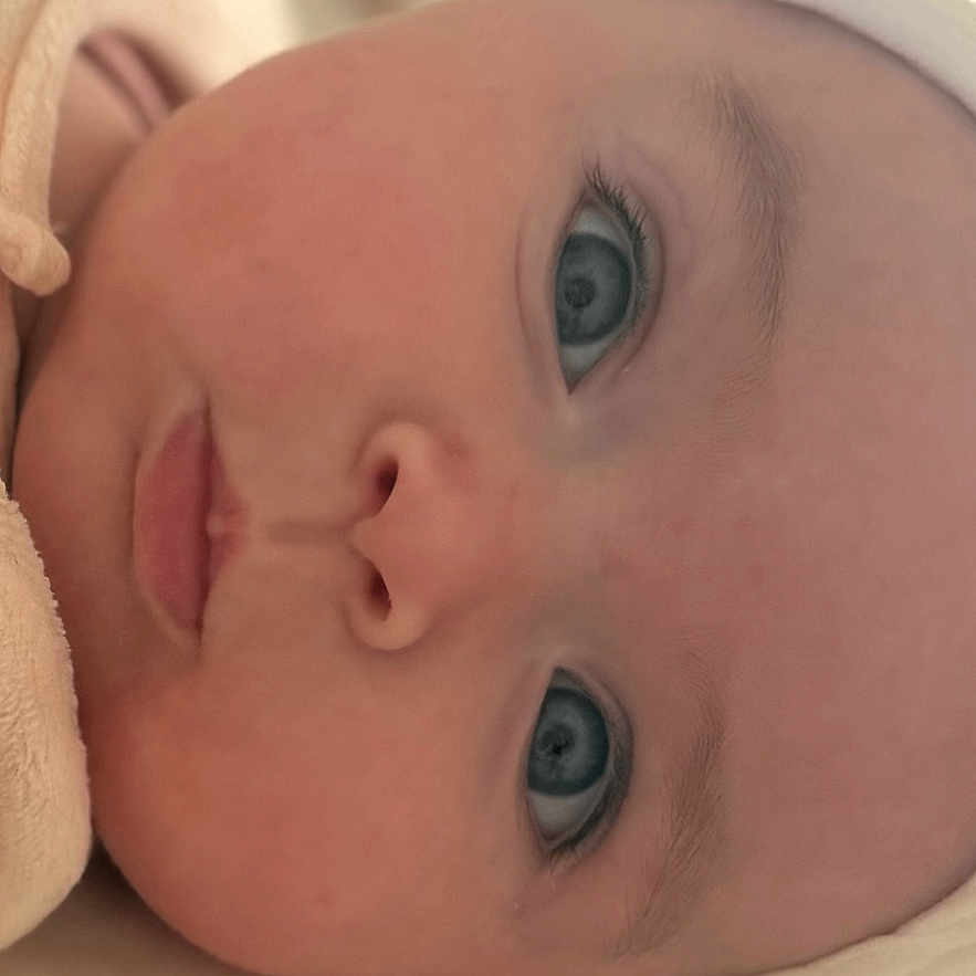 Cassie participe au concours pour gagner de l'argent avec cette photo : baby, infant, face, blue_eyes, close_up, headwear, skin, portrait, lying_down, sleepwear, newborn, child, soft_light, indoor, expressionless, cute, human, person, head, background_blur