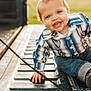 toddler, child, smiling, blue_eyes, jeans, shirt, outdoor, sunlight, happy, baby, portrait, sitting, hand, metal_surface, patterned_clothing, cute, casual_wear, closeup, daylight, playful