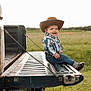 toddler, cowboy_hat, jeans, boots, pickup_truck, tailgate, field, grass, smile, child, outdoor, sunlight, belt_buckle, shirt, rural, nature, happy, portrait, casual, sitting