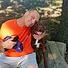 affection, canine, dog, grass, happy, human, leash, man, nature, orange_shirt, outdoor, park, pet, shoes, shorts, sitting, smile, stone, sunlight, tree