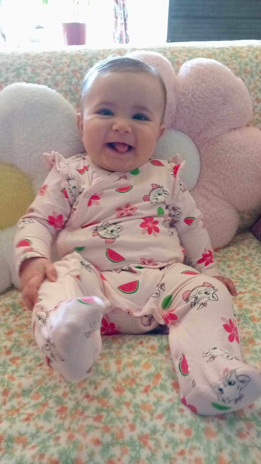 Myana participe au concours pour gagner de l'argent avec cette photo : baby, child, infant, smiling, pink_onesie, watermelon_print, flower_print, couch, floral_pattern, cushion, soft_toys, happy, tongue_out, indoors, cute, sitting, person, young_child, playful, cozy