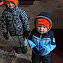 Waylon is registered to the contest to win money with this photo: child, toddler, winter_clothing, hat, gloves, jacket, indoor, carpet, floor, cold_weather, orange_hat, blue_jacket, checkered_jacket, boots, person, standing, home, laundry_machine, dog, shadow