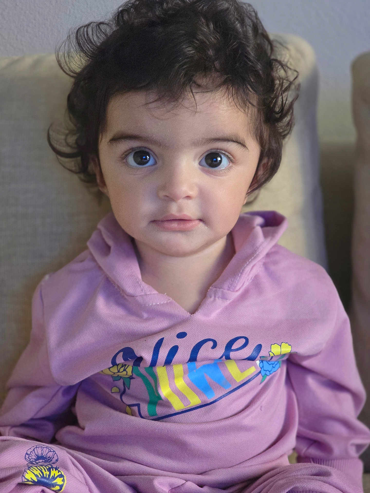 Haya is registered to the contest to win money with this photo: child, toddler, curly_hair, purple_hoodie, sitting, couch, indoor, face, large_eyes, portrait, clothing, person, young_child, casual, expression, comfortable, home, cute, looking_at_camera, cozy