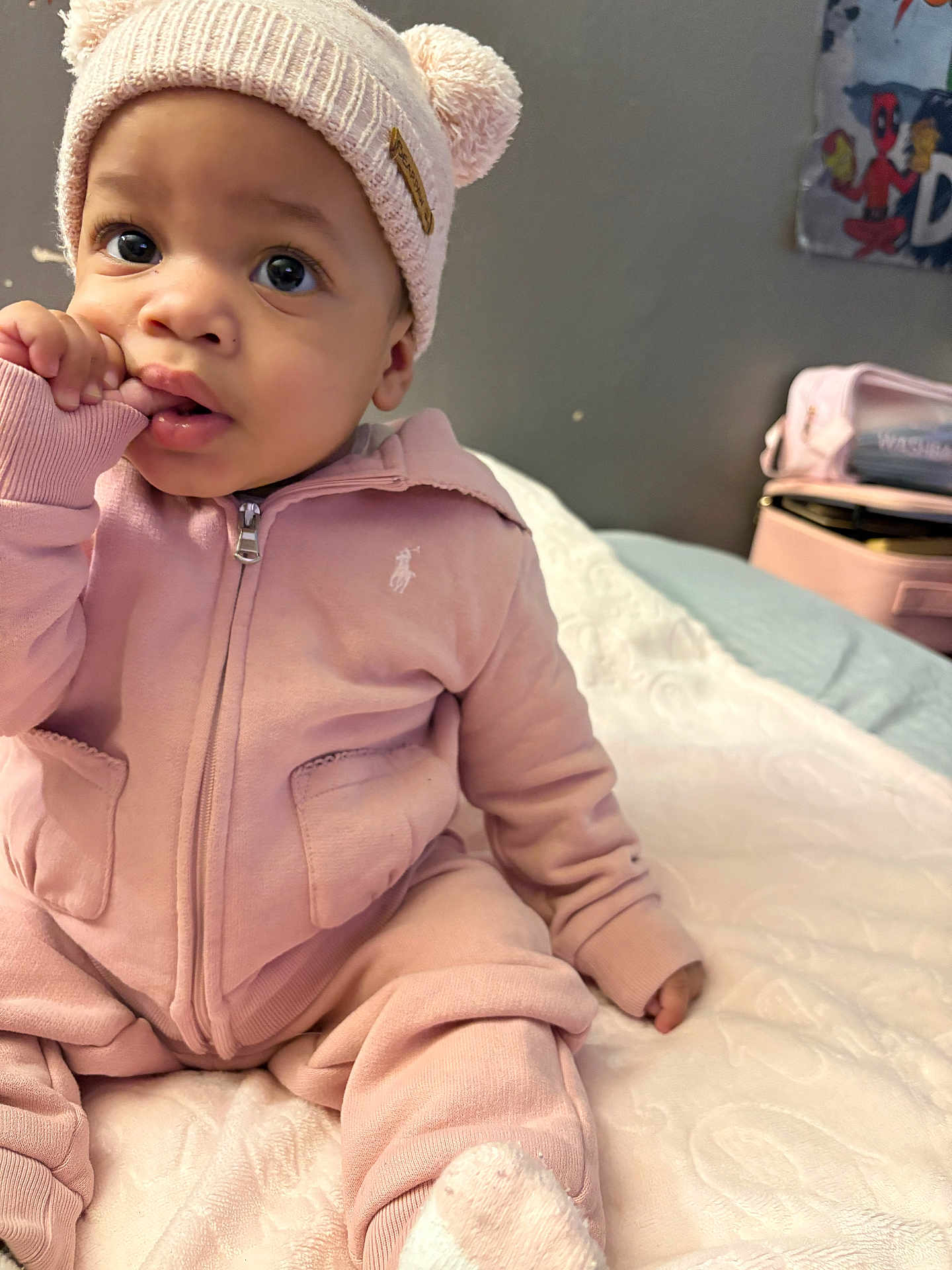 Jenaiyah is registered to the contest to win money with this photo: baby, child, pink_clothing, hat, knit_hat, bear_ears, blanket, indoor, bed, curious, face, person, cute, sitting, portrait, cozy, soft_texture, expression, finger_in_mouth, young_child