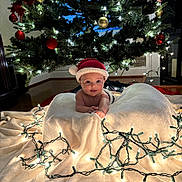 Grace joined the competition — help win amazing prizes! baby, santa_hat, blanket, christmas_tree, christmas_lights, ornaments, holiday, indoor, festive, decorations, toddler, face, smiling, candle_lights, floor, warm_lighting, cute, festivity, celebration, christmas