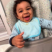 Aiden joined the competition — help win amazing prizes! baby, bib, child, clothing, curly_hair, cute, expression, face, furniture, happy, high_chair, indoor, infant, joyful, messy, person, seat, smiling, thumbs_up, tray