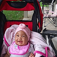 Kayzlee is registered to the contest to win money with this photo: baby, stroller, pink_hat, pink_bib, blanket, smiling, happy, outdoor, porch, grill, chair, baby_clothing, infant, child, person, seat, fabric, blanket_edge, daylight, relaxing