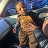 baby, child, car_interior, overalls, brown_clothing, hand, adult, sunlight, window, car_seat, curious_expression, casual, daylight, trees, parked_cars, footwear, person, portrait, indoor, support