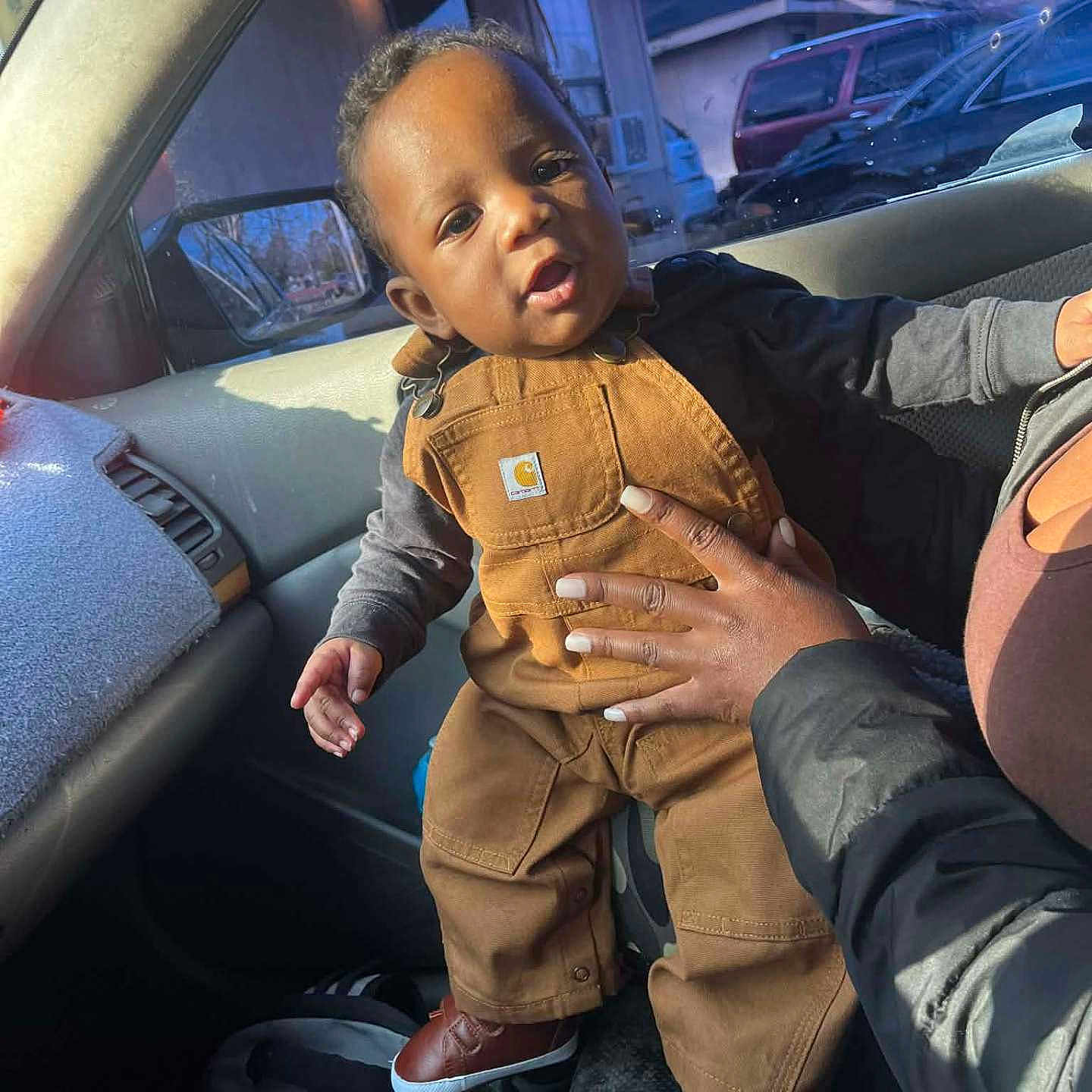 Trustin is registered to the contest to win money with this photo: baby, child, car_interior, overalls, brown_clothing, hand, adult, sunlight, window, car_seat, curious_expression, casual, daylight, trees, parked_cars, footwear, person, portrait, indoor, support