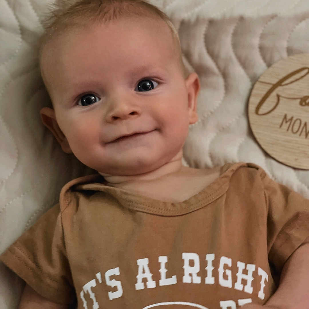 Weston joined the competition — help win amazing prizes! baby, infant, smiling, face, portrait, clothing, brown_shirt, quilted_blanket, lying_down, cute, young_child, happy, indoors, human, child, head, skin, eyes, hand, expression