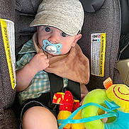 Weston is registered to the contest to win money with this photo: baby, child, car_seat, pacifier, cap, plush_toy, blue_eyes, bib, checkered_shirt, seatbelt, indoor, cute, infant, toy, colorful, soft_toy, young_child, sitting, casual_clothing, car_interior
