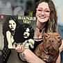 album_cover, black_top, brown_dog, casual_clothing, curly_hair, dog, face, glasses, hand, holding, indoor, necklace, pet, portrait, red_collar, sitting, smiling, tattoo, vinyl_record, woman