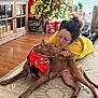 dog, woman, yellow_hoodie, glasses, christmas_tree, holiday_decorations, gift_boxes, wooden_floor, rug, indoor, pet, affection, bandana, red_bandana, festive, holiday_season, living_room, casual_clothing, glasses_woman, christmas