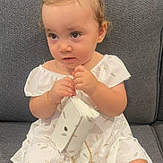 Amelia participe au concours pour gagner de l'argent avec cette photo : casual, child, curious_expression, curly_hair, cute, fashion, girl, gold_chain, gold_feathers, gray_couch, hair_buns, indoors, portrait, purse, sitting, small_hands, soft_lighting, toddler, white_dress, young_child