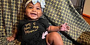 Nevaeh is registered to the contest to win money with this photo: baby, smiling, child, headband, bow, clothing, black_outfit, blanket, pattern, chair, indoors, happy, person, baby_leg, baby_foot, cute, infant, sitting, room, fabric