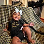 baby, smiling, child, headband, bow, clothing, black_outfit, blanket, pattern, chair, indoors, happy, person, baby_leg, baby_foot, cute, infant, sitting, room, fabric
