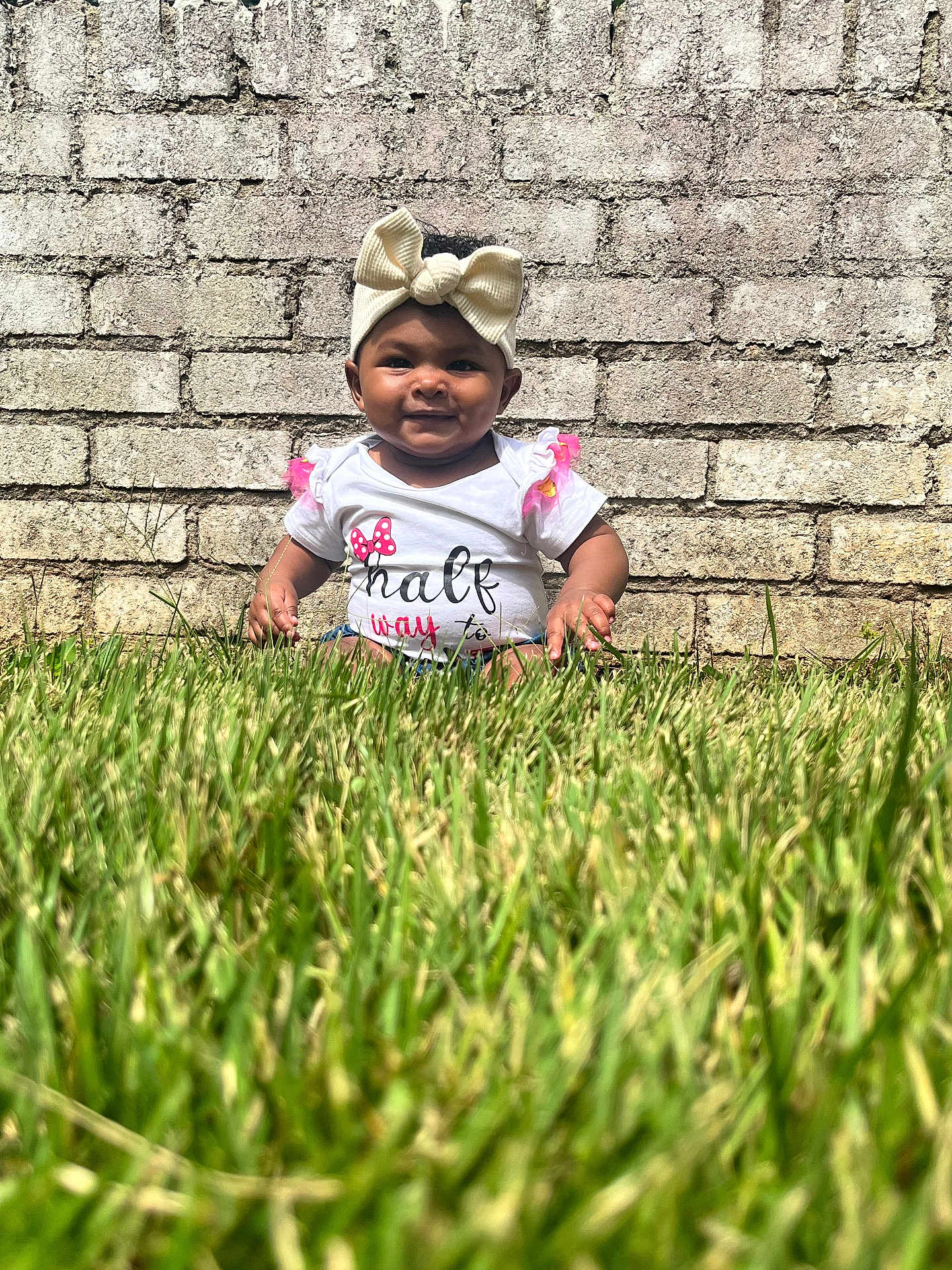 Nevaeh is registered to the contest to win money with this photo: baby, child, grass, brick_wall, headband, bow, white_shirt, outdoor, smile, happy, sunny, infant, cute, sitting, greenery, nature, portrait, person, daylight, casual
