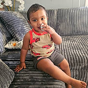 Zaden Ramjohan joined the competition — help win amazing prizes! toddler, child, sitting, sofa, couch, smiling, portrait, indoor, living_room, snack, food, hand, pajamas, car_pattern, gray, cushion, flowers, radiator, cozy, photography