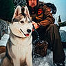 husky, dog, snow, man, woman, winter_clothing, outdoor, trees, graffiti, leash, fur_hat, red_hood, cold_weather, sitting, portrait, nature, snowy_ground, smiling_dog, people, daylight