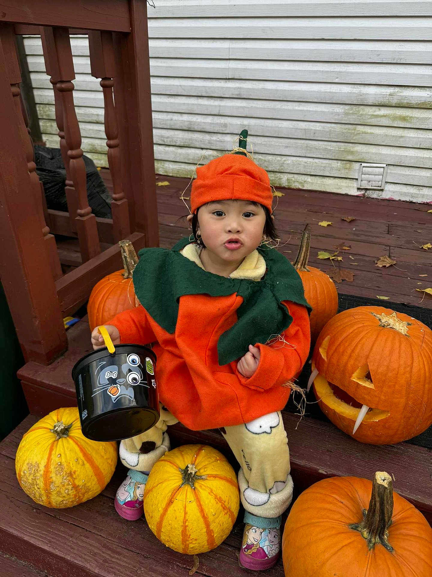 Khai My joined the competition — help win amazing prizes! child, toddler, pumpkin_costume, halloween, pumpkins, bucket, orange, green, wooden_steps, fall, festive, cute, outdoor, seasonal, costume, decorations, playful, expression, clothing, holiday