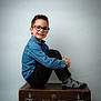 child, boy, glasses, denim_shirt, black_pants, socks, dinosaur_pattern, vintage_suitcase, smile, portrait, studio, seated, indoor, happy, casual_clothing, short_hair, light_background, fashion, cute, young