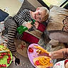 child, toddler, pajamas, striped_clothing, couch, pillows, hot_dog, ketchup, cheese, plate, bottle, window, rug, penguin_pattern, snowflake_pattern, indoor, smiling, food, person, tattoo