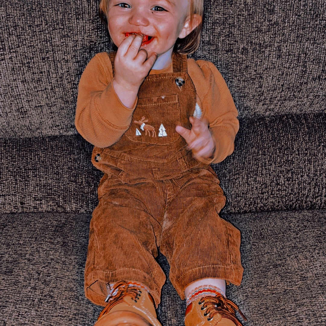 Rodger joined the competition — help win amazing prizes! toddler, child, smiling, couch, brown_overalls, boots, sitting, happy, indoors, face, hair, cute, playful, clothing, person, young_child, texture, cozy, casual, portrait