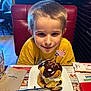 child, boy, dessert, chocolate, ice_cream, plate, spoon, table, restaurant, booth, napkin, glass, yellow_shirt, happy, food, indoor, flag, sweet, smile, person