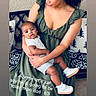 woman, baby, child, green_dress, white_shoes, striped_shorts, bench, pillow, indoor, floor_tiles, curly_hair, holding, portrait, family, love, casual_clothing, text_overlay, smiling, affection, seated