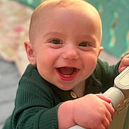 Bryce is registered to the contest to win money with this photo: baby, smiling, happy, child, face, hand, green_clothing, closeup, indoor, infant, cute, portrait, young_child, holding, railing, joy, bright_eyes, skin, expression, warm_lighting
